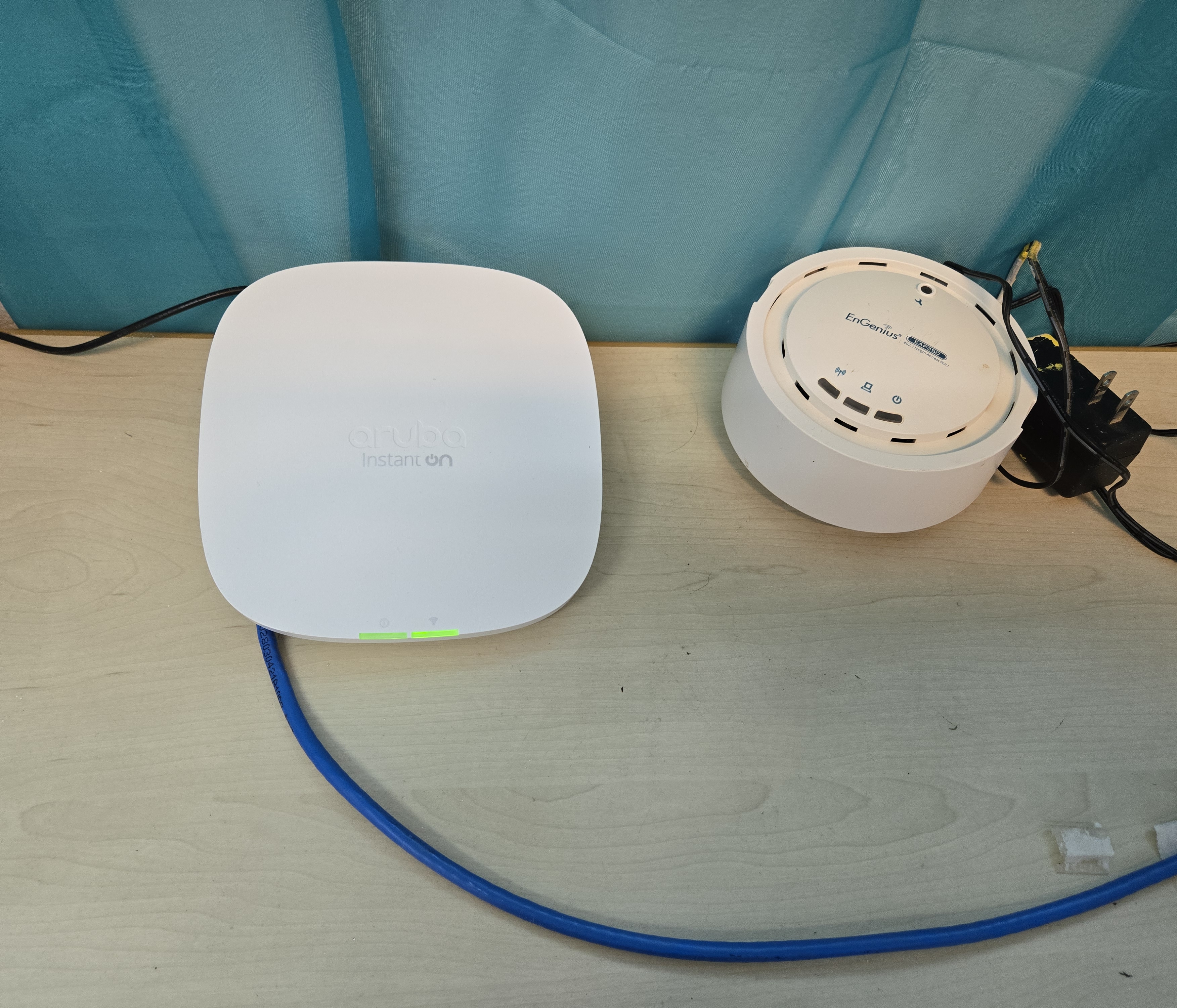 Aruba AP22 Instant On (izquierda, LEDs verdes activos) junto al equipo EnGenius reemplazado (derecha). Cable LAN azul conectado.